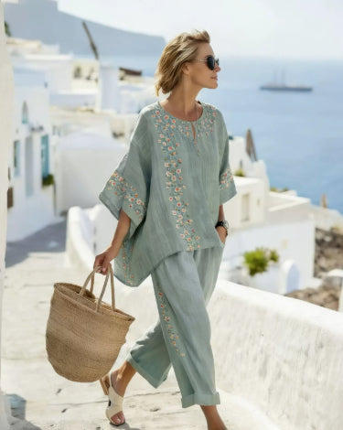 Woman in light blue outfit with floral patterns walking on a sunlit street with white buildings and blue sea.