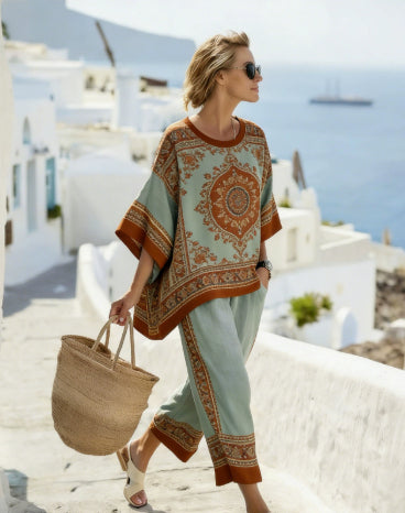 Woman in a patterned outfit with a straw bag standing in a scenic location with white buildings and blue sky.